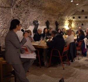 Una relatrice con microfono presenta una degustazione guidata agli ospiti seduti a una lunga tavolata, in una cantina sotterranea con volta in pietra e sculture alle pareti, a Testaccio, Roma.
