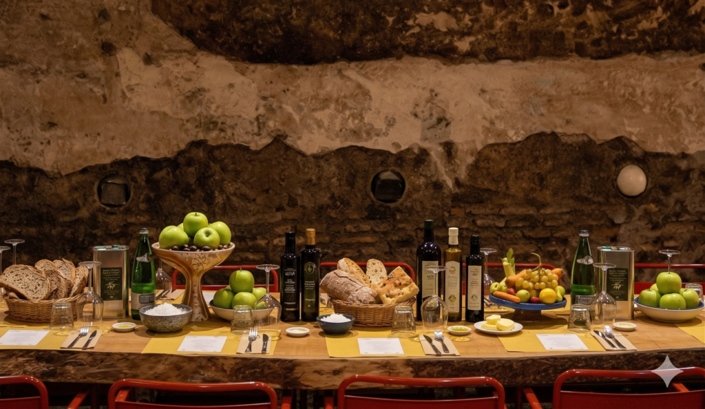 Tavolata apparecchiata per una degustazione di olio extravergine d’oliva, con pane, mele verdi e bottiglie d’olio, in una sala sotterranea con muro antico a Testaccio, Roma.