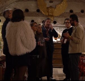 Gruppo di ospiti in piedi con i calici in mano ascolta la presentazione di un vino rosato, in una cantina sotterranea con una scultura dorata ad ali spiegate alla parete, a Testaccio, Roma.