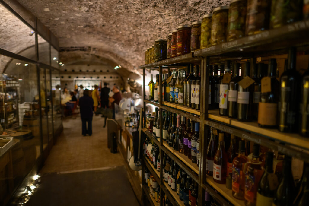Wood and iron shelves stocked with white, red and rosé wine bottles and jars of Italian preserves in olive oil, along the corridor of an underground stone-vaulted wine cellar in Testaccio, Rome.