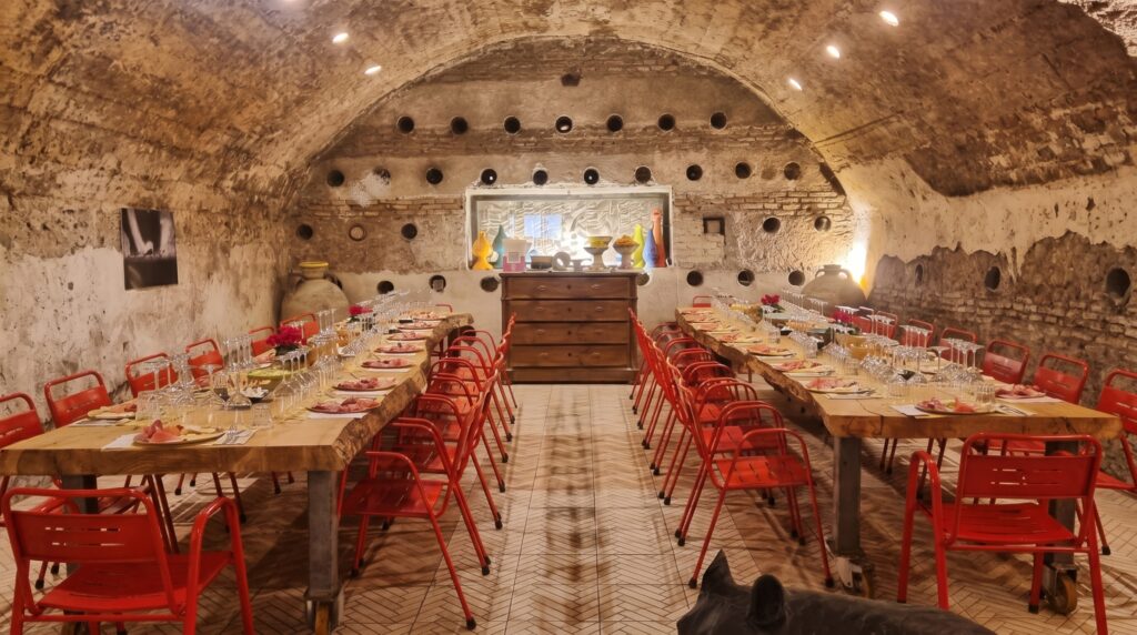 Cantina sotterranea con volta a botte in pietra e mattoni, due lunghe tavolate in legno apparecchiate con piatti di salumi e formaggi, calici e sedie rosse, anfore e vasi in ceramica colorata, a Testaccio, Roma.