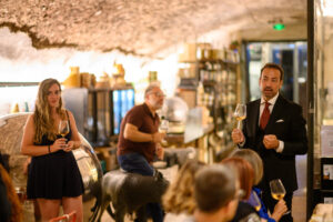 Sommelier in giacca e cravatta presenta un calice di vino bianco agli ospiti durante una degustazione, in una cantina sotterranea con volta in pietra e scaffali di prodotti tipici, a Testaccio, Roma.