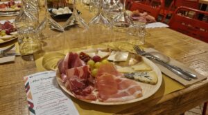 Piatto di degustazione con prosciutto crudo, capocollo, salame, formaggi stagionati ed erborinati e acini d’uva, su una tavola in legno apparecchiata con calici e menu, a Testaccio, Roma.
