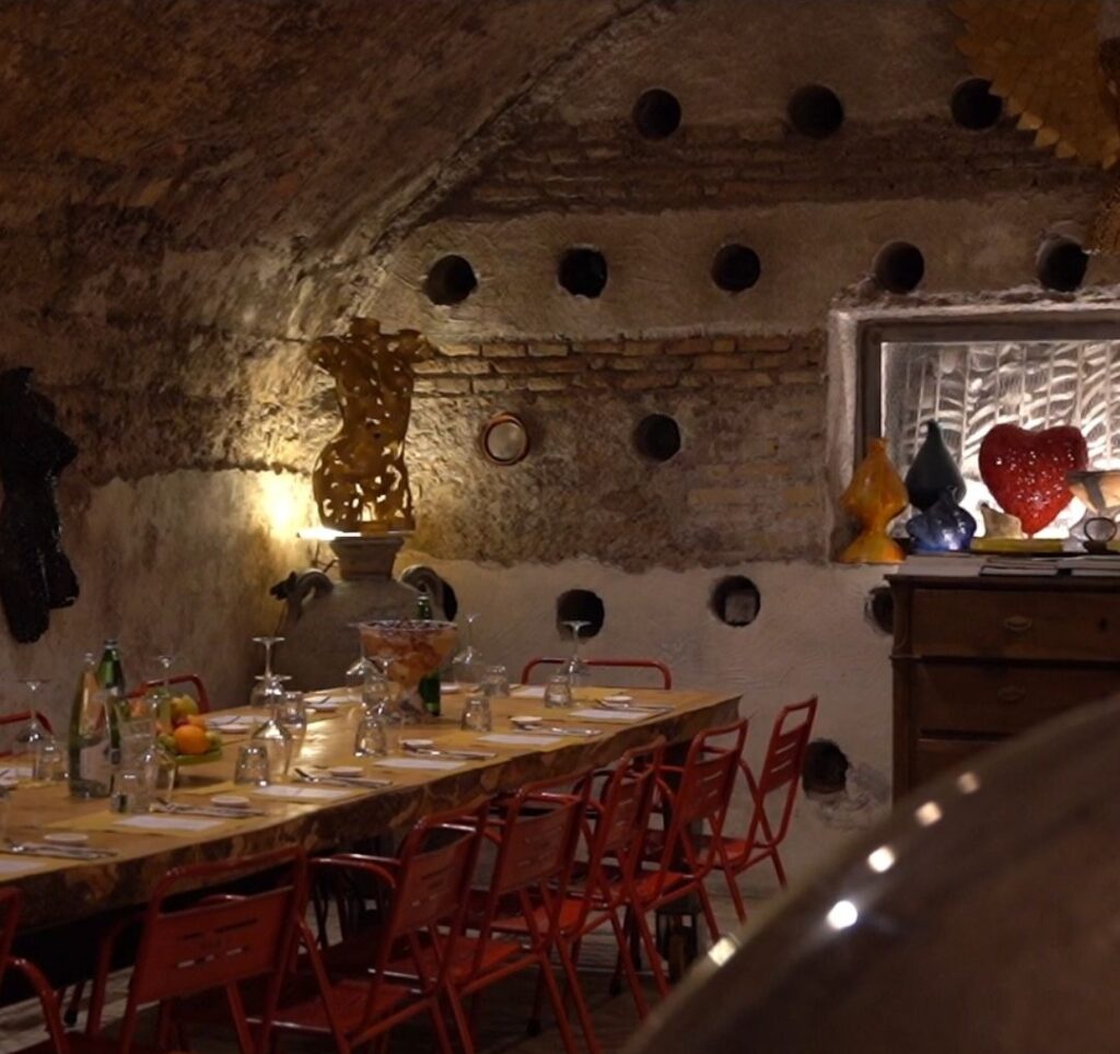 Long wooden table set with wine glasses, water bottles and fresh fruit, with red chairs, in an underground stone-vaulted wine cellar with round wall niches, golden sculptures and colourful glass art in Taste'accio, Rome.
